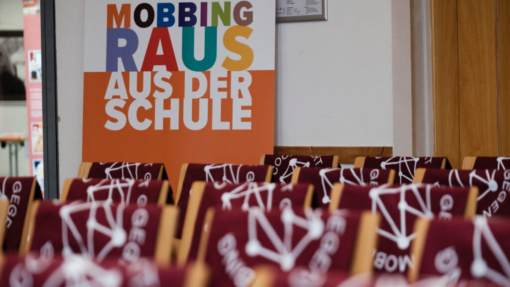 Vorne leere Stuhlreihen, hinten Schriftzug Mobbing raus aus der Schule