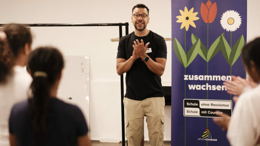 Workshopleiter neben einem Roll-up "Zusammenwachsen" und Hinterköpfe von Kindern, die ihm zuhören.