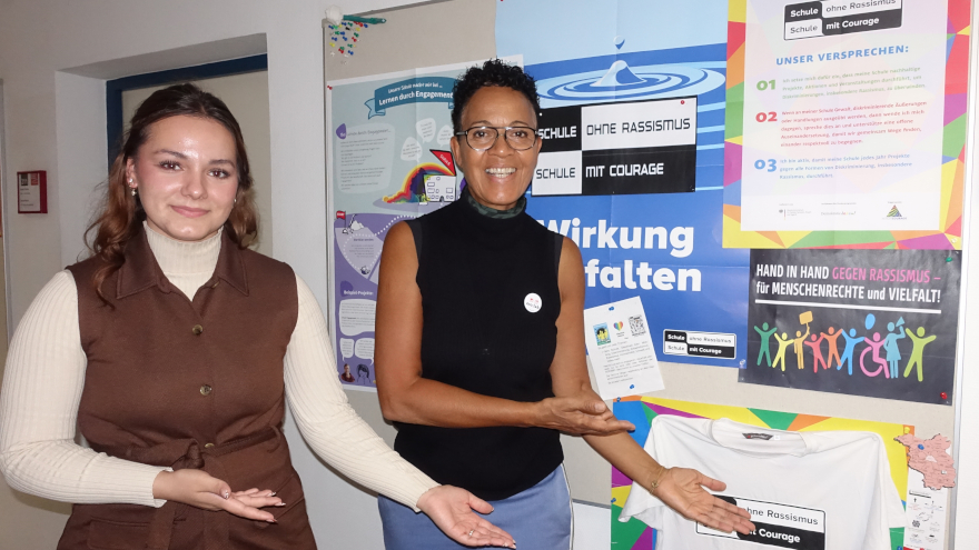 Zwei Frauen zeigen auf eine Pinnwand mit Postern von Schule ohne Rassismus - Schule mit Courage
